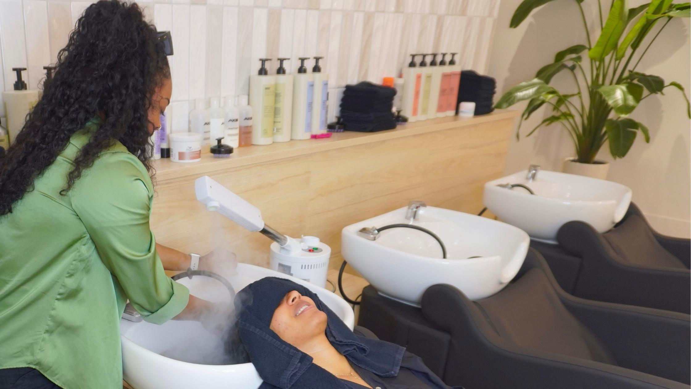 A woman with curly hair braids is having her hair washed at a salon, relaxing in a salon chair.