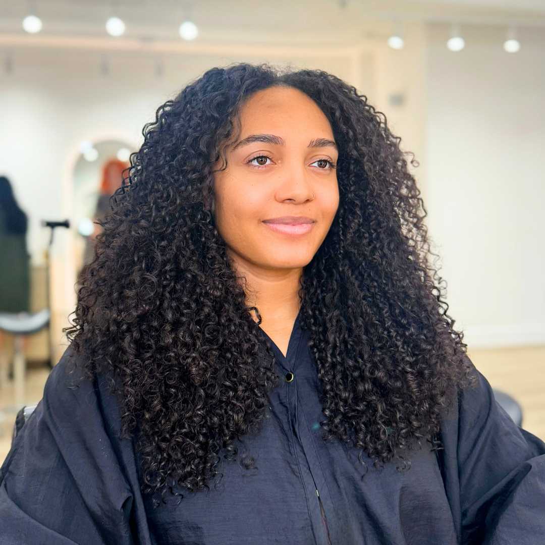 A woman with long, curly hair seated in a salon, ready for a curly hair treatment.