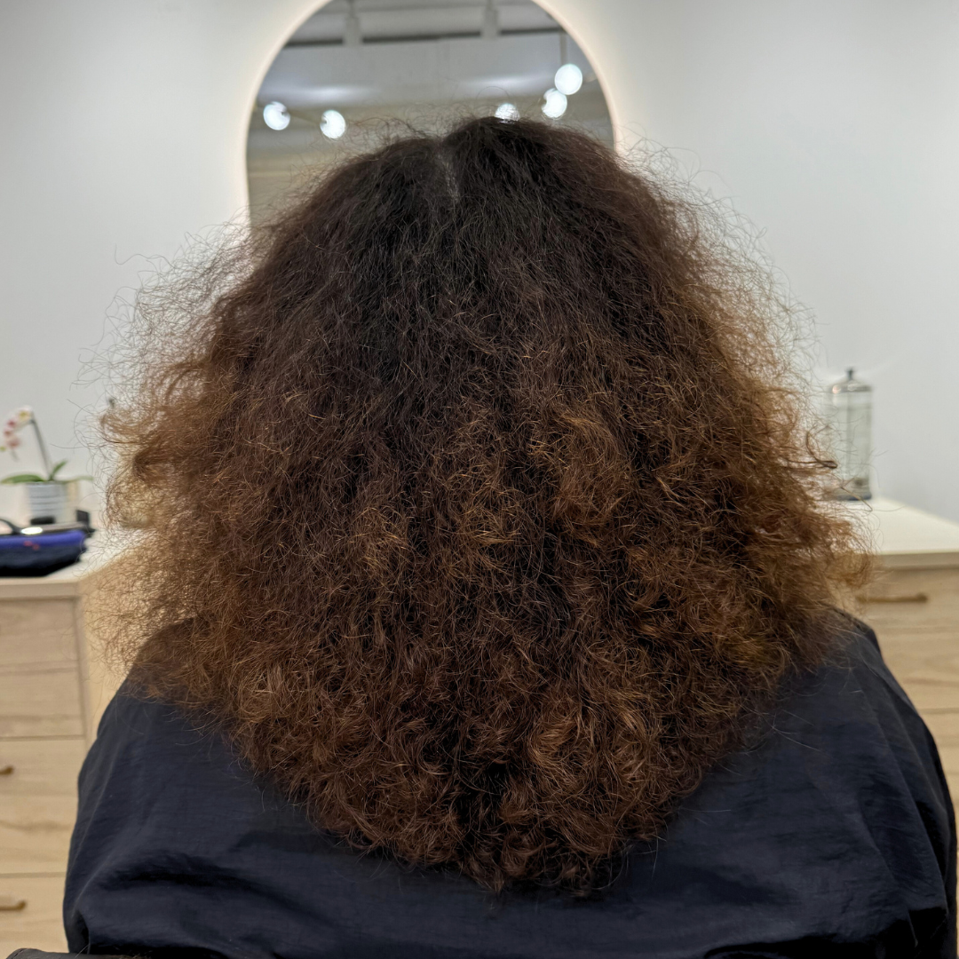 A woman with long curly hair styled in braids sits comfortably in a chair, looking relaxed and content.